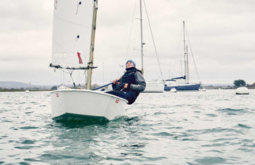 A young sailor sailing an Optimist dinghy, wearing a grey Pro Lite Aquafleece top, junior coastal hi-fits, a black Race Armour buoyancy aid, and an Aquafleece beanie. The sailor is smiling while navigating calm waters, with moored yachts and a cloudy sky in the background.
