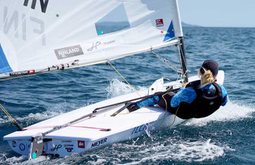 A sailor hiking out on a Laser dinghy in moderate wind, wearing a women's Thermaflex Longjohn and Top in Seagrass, along with a black Race Armour buoyancy aid and a cap. The boat features Finnish team branding and multiple sponsor logos, sailing briskly across open water on a sunny day.