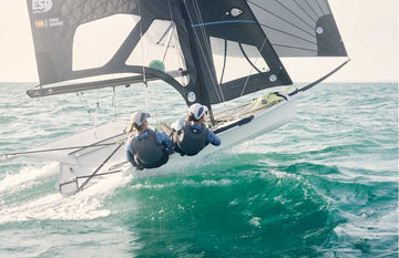 Two women sailors are actively sailing a 49er dinghy. They are both wearing Rooster Women's Thermaflex Longjohn wetsuits and matching tops in Seagrass, along with grey race bibs. On their heads, they wear white Rooster Aeromesh 2.0 caps. The scene captures their focus and teamwork as they navigate the water.