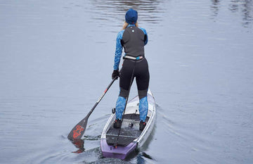 A woman paddleboards across calm water, wearing the Rooster Women's Thermaflex Longjohn and top in Seagrass. She has on low-cut boots, Pro Race gloves, and a blue and black Supertherm beanie, staying warm and comfortable as she paddles